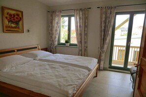 Een slaapkamer met een tweepersoonsbed, een raam met gordijnen, een potplant en uitzicht op een balkon.