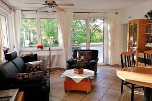 A living room with a sofa, armchair, coffee table, and a dining table. There is a radiator, a ceiling fan, and a bookshelf. - Carpe Diem Lake Park (Seebad Heringsdorf)