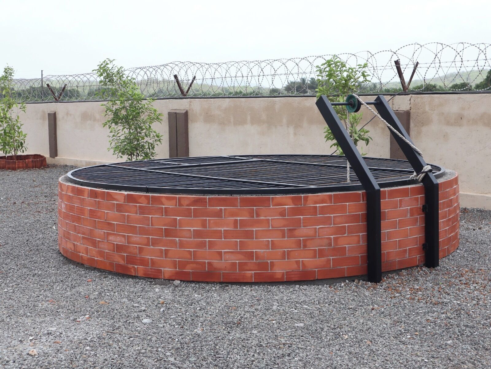 A circular brick structure with a metal grill, surrounded by gravel, with a barbed wire fence in the background.