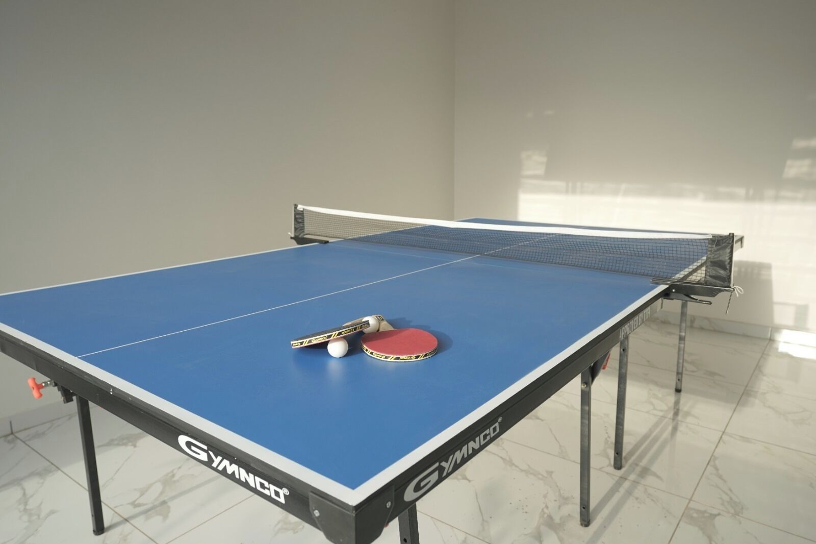 A table tennis table with a net, a red table tennis paddle, and two balls.