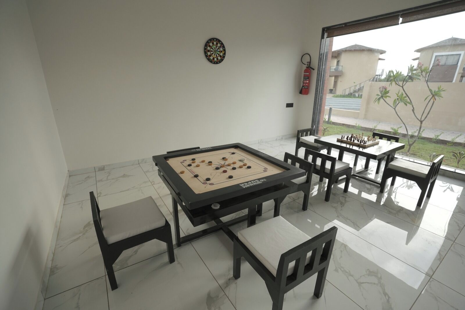 Une table de billard avec des chaises, un jeu de fléchettes et un extincteur dans une pièce donnant sur l’extérieur.