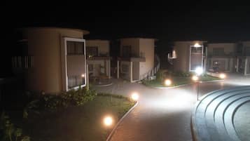 Vista nocturna de un hotel con varias construcciones, senderos iluminados y pequeños jardines.