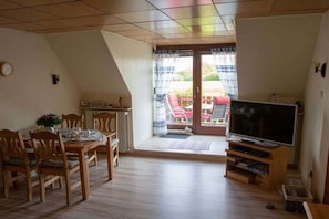 A dining area with a table and chairs, a television on a wooden stand, and a sliding glass door leading to an outdoor seating area. - "At Hahnheide" || Linau (Linau)