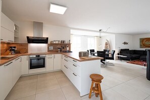 A modern kitchen with a central island, white cabinetry, and a dining area with a black sofa and chairs. - Holiday home with 2 bedrooms (18445 Kramerhof, Deutschland)