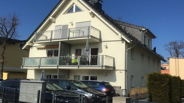 Un edifici residencial de dues plantes amb un balcó al pis superior, una zona d'aparcament tancada amb cotxes i un cel serè.