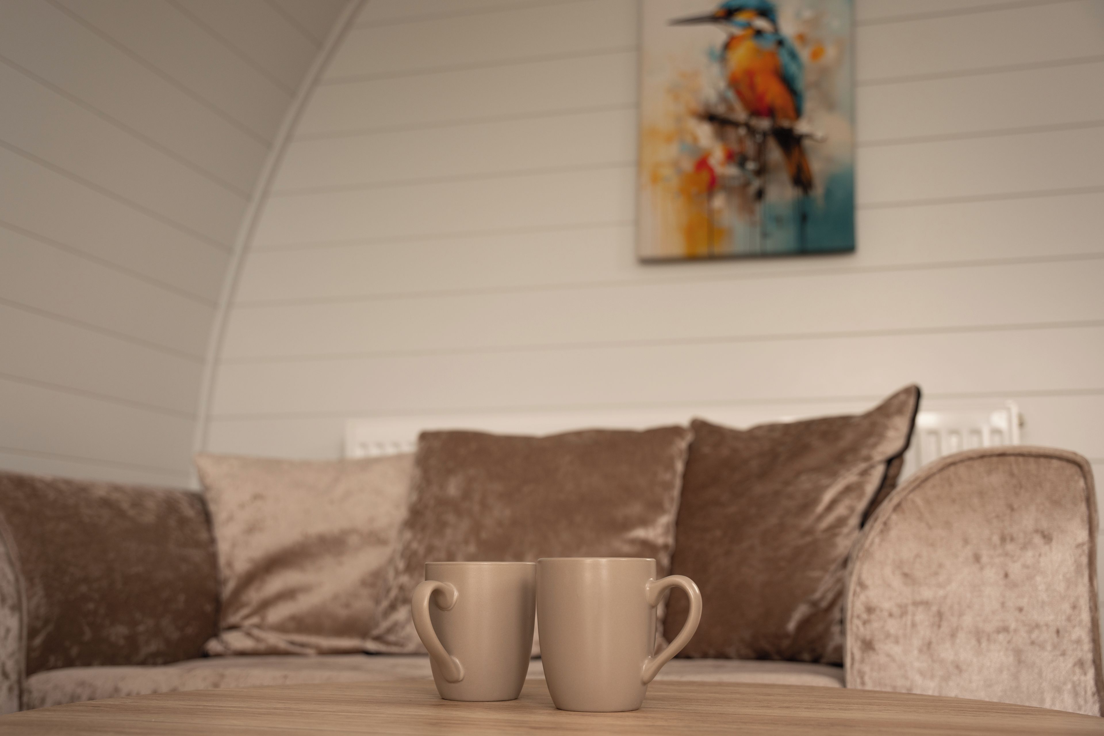 A room with a wooden table, two beige mugs, a beige sofa, and a painting of a bird on the wall.