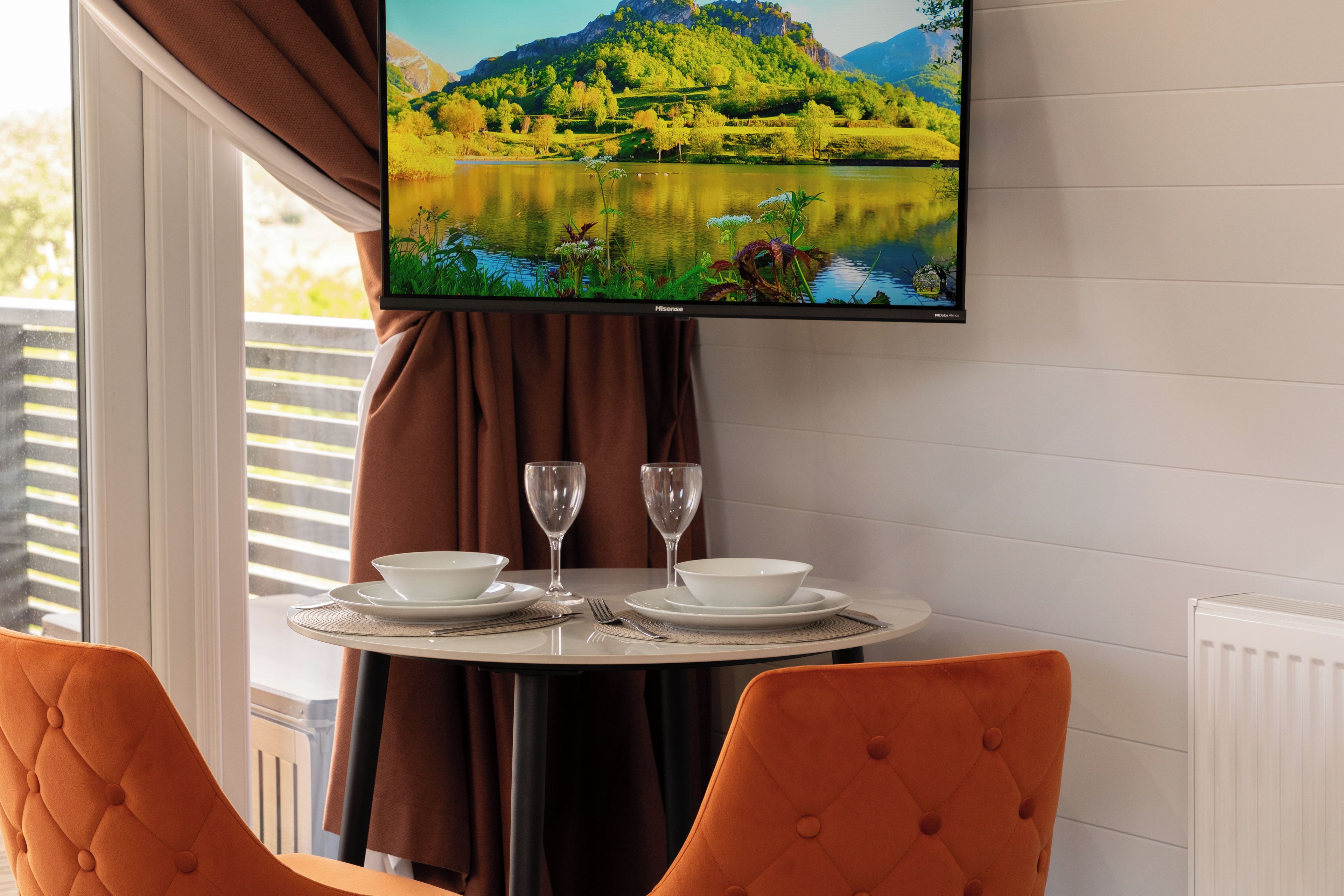 A dining area with a table set for two, a television mounted above displaying a scenic landscape, and a window with curtains.