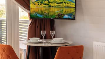 A dining area with a table set for two, a television mounted above displaying a scenic landscape, and a window with curtains.