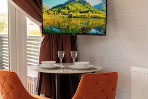A dining area with a table set for two, a television mounted above displaying a scenic landscape, and a window with curtains.
