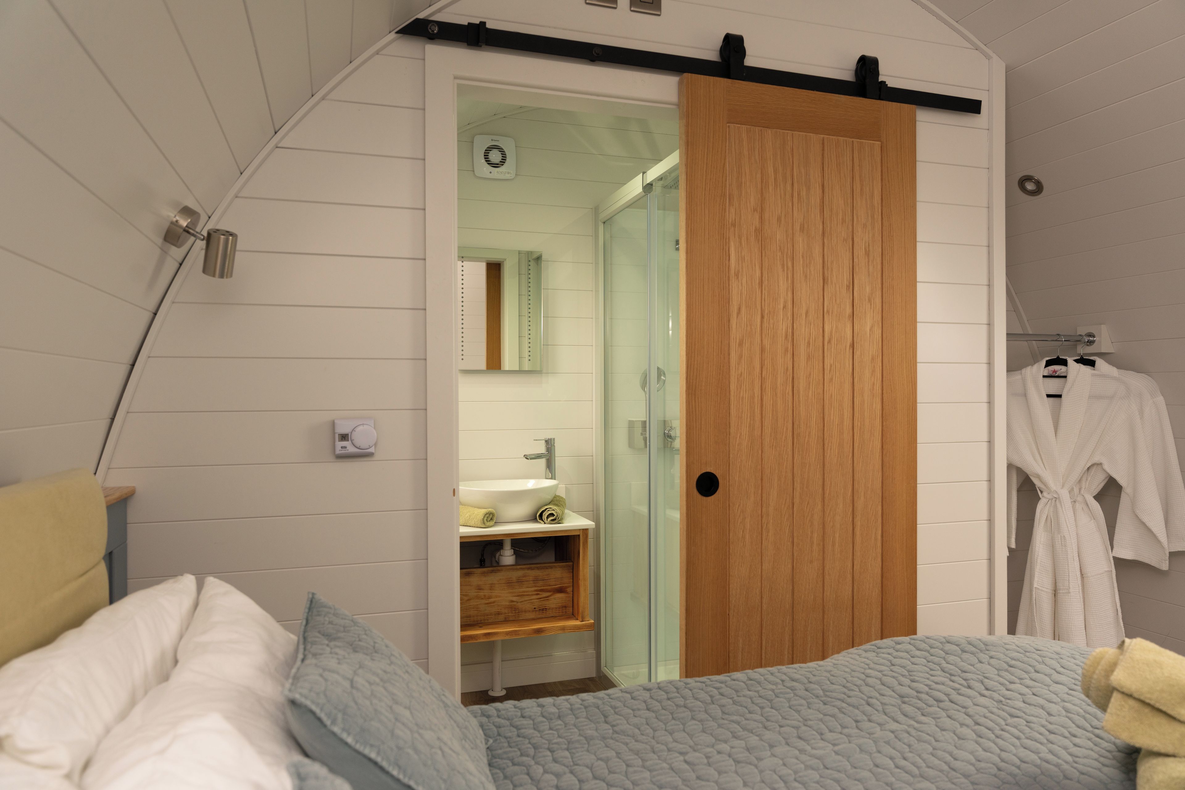 A bedroom with a bed, a wooden wardrobe, a bathroom with a sink and mirror, and a robe hanging on a rack.
