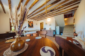 Interior rústic amb bigues de fusta a la vista, zona de menjador amb taula i cadires de fusta, sofà i zona de cuina amb nevera i armaris.