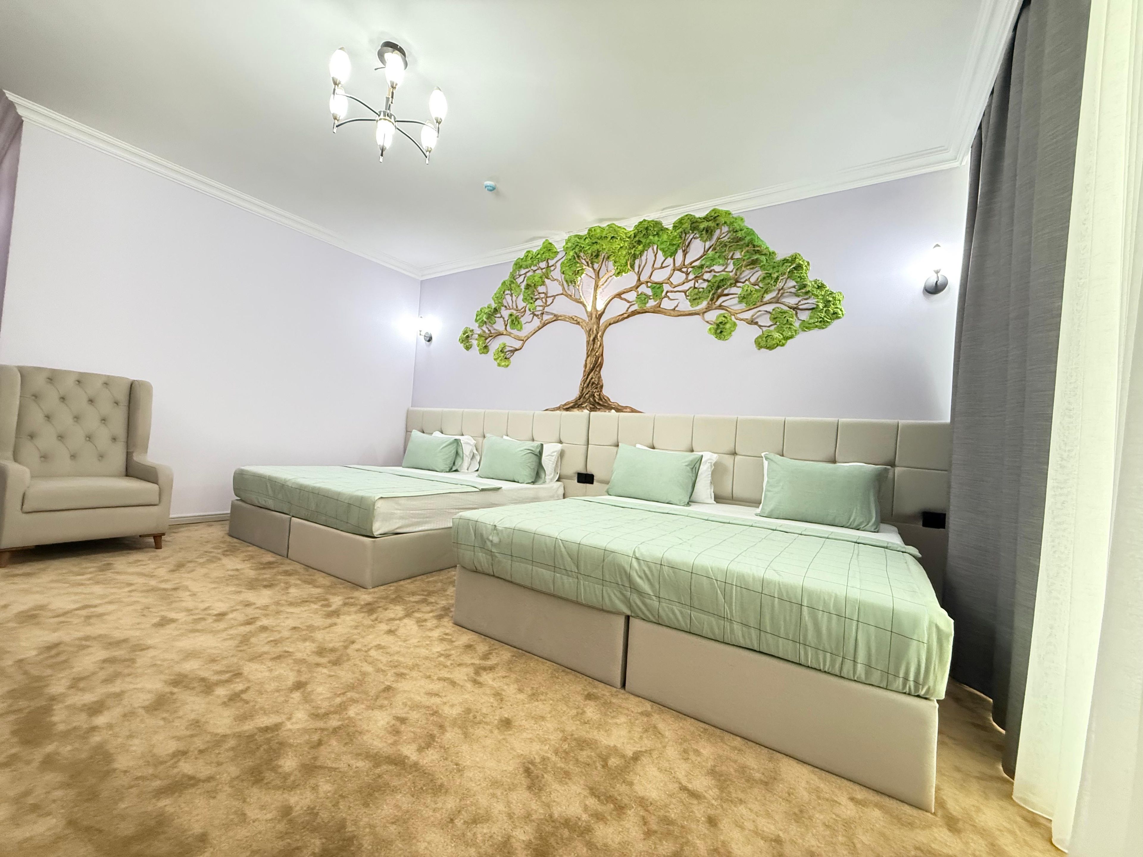 A bedroom with two beds, a chandelier, a tree mural, and a beige armchair.