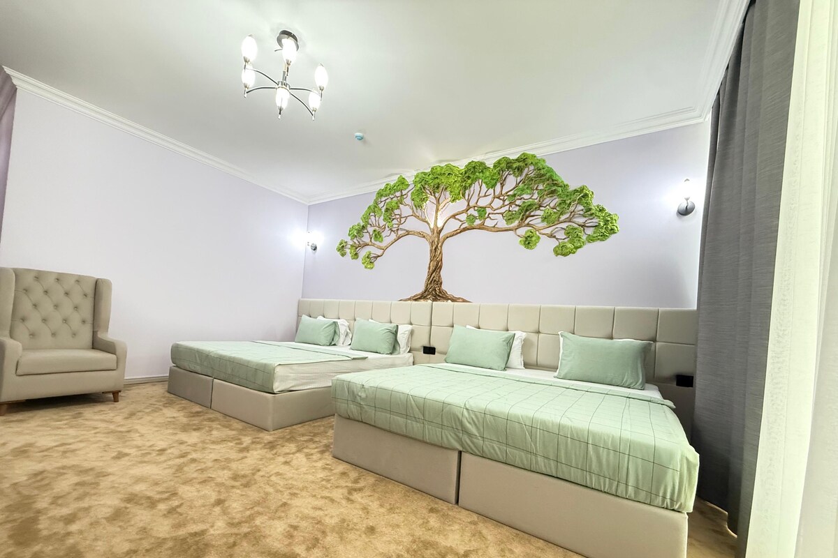 A bedroom with two beds, a chandelier, a tree mural, and a beige armchair.