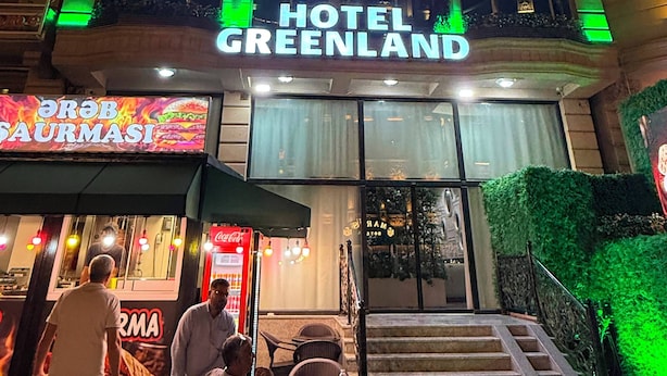 Un hotel llamado 'HOTEL GREENLAND' con un letrero llamativo, una escalera que conduce a la entrada y un restaurante llamado 'GORB SAURMASI' visible al frente.