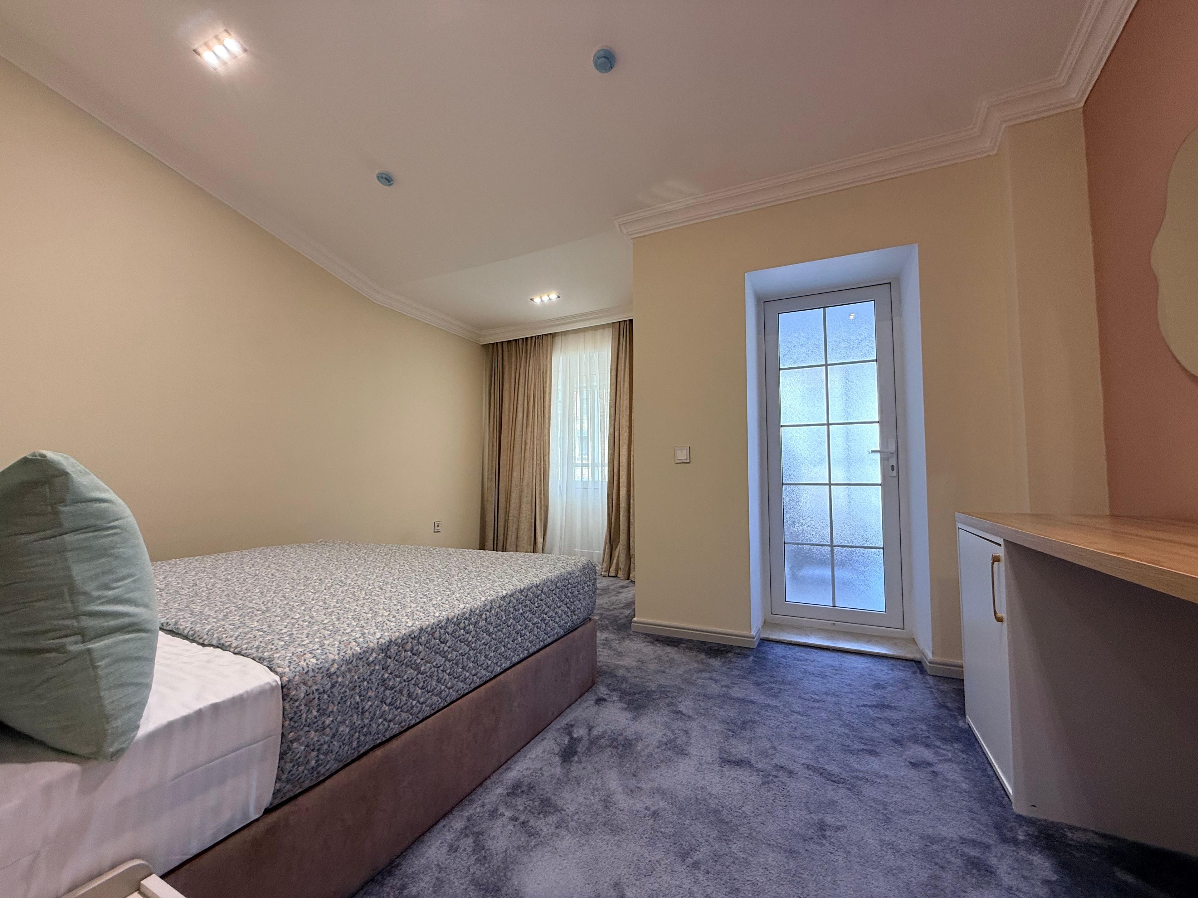 A bedroom with a bed, a desk, a door, and a window with curtains.