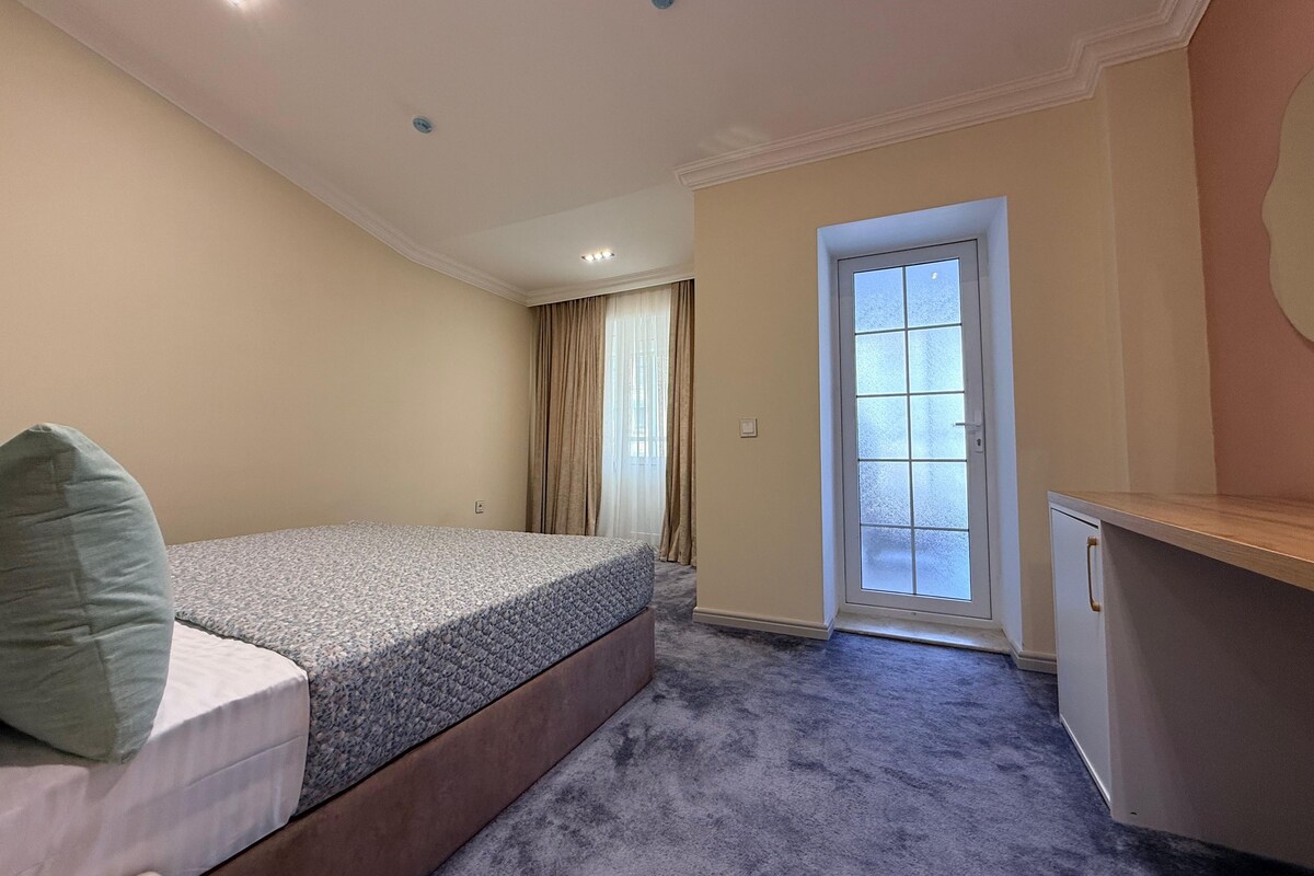 A bedroom with a bed, a desk, a door, and a window with curtains.