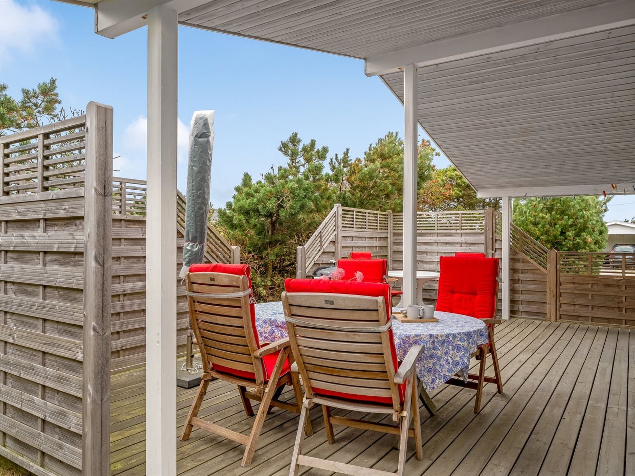 Une terrasse extérieure avec un sol en bois, deux chaises en bois dotées de coussins rouges, une petite table ronde recouverte d’une nappe à motifs floraux et un espace abrité avec une rambarde en bois.