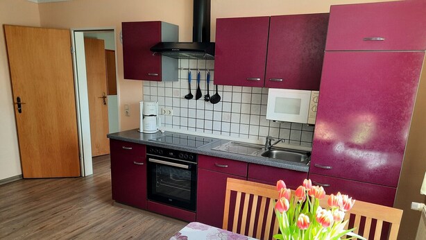 Eine kompakte Küche mit Mikrowelle, Herd und Spüle. Ein Esstisch mit Blumenvase und Korb sind ebenfalls vorhanden.