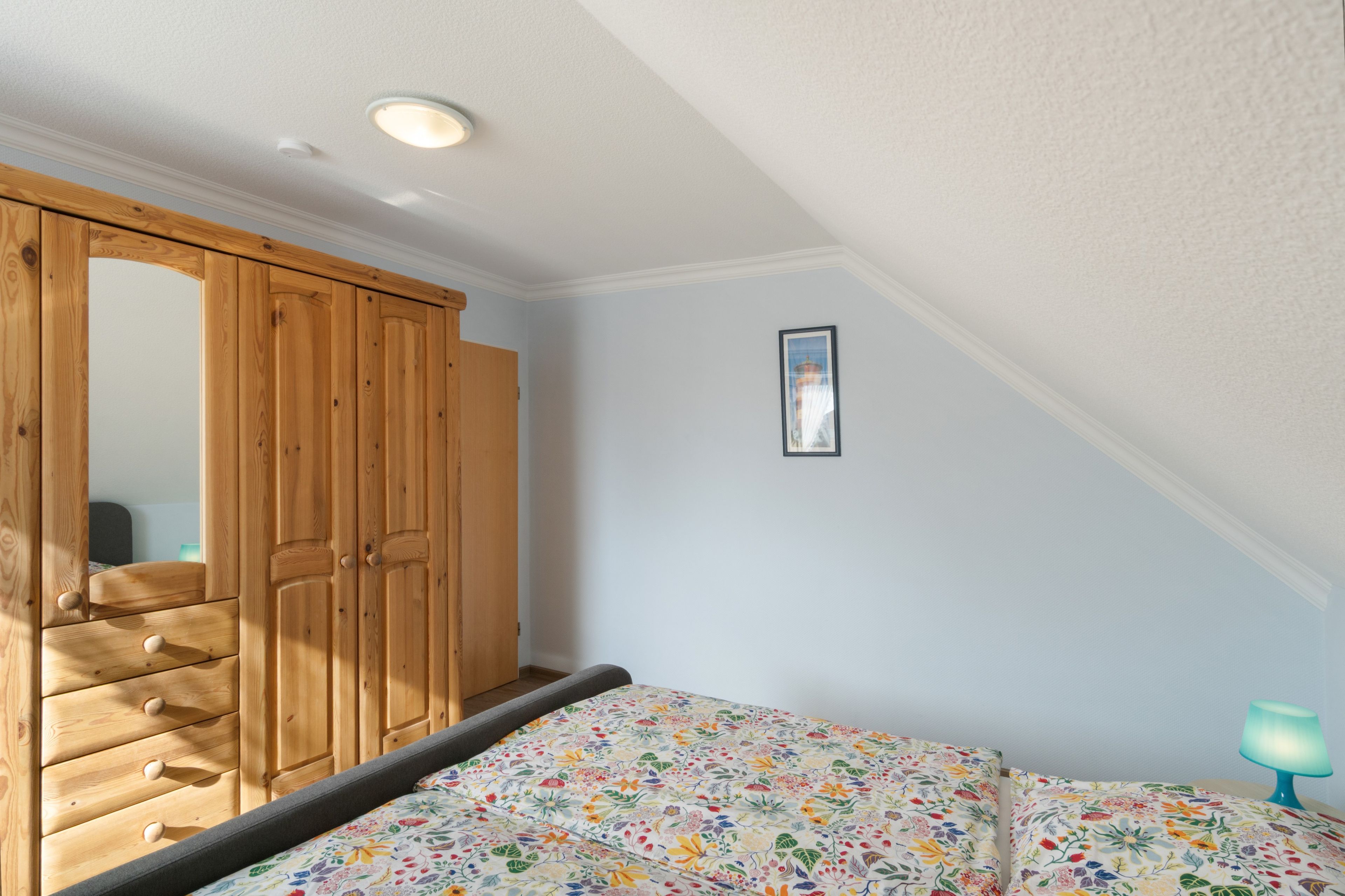 Een slaapkamer met een houten garderobe, een bed met een bloemenprint, een lijst met een afbeelding aan de muur en een nachtkastje met een lamp.