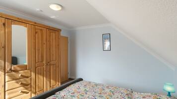 Een slaapkamer met een houten garderobe, een bed met een bloemenprint, een lijst met een afbeelding aan de muur en een nachtkastje met een lamp.