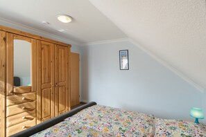 Een slaapkamer met een houten garderobe, een bed met een bloemenprint, een lijst met een afbeelding aan de muur en een nachtkastje met een lamp.