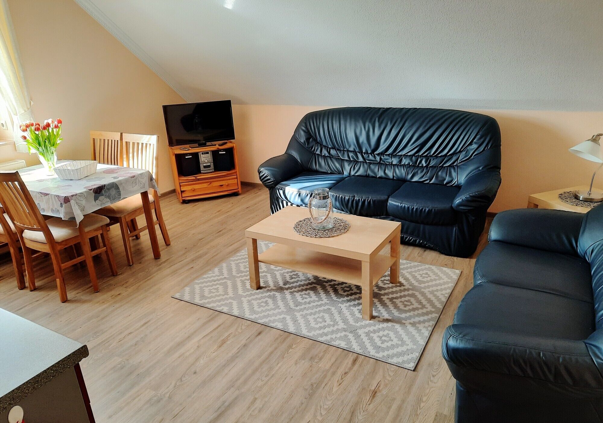 Ein Wohnzimmer mit einem schwarzen Ledersofa, einem Holztisch, einem Esstisch mit Stühlen und einer TV-Konsole.