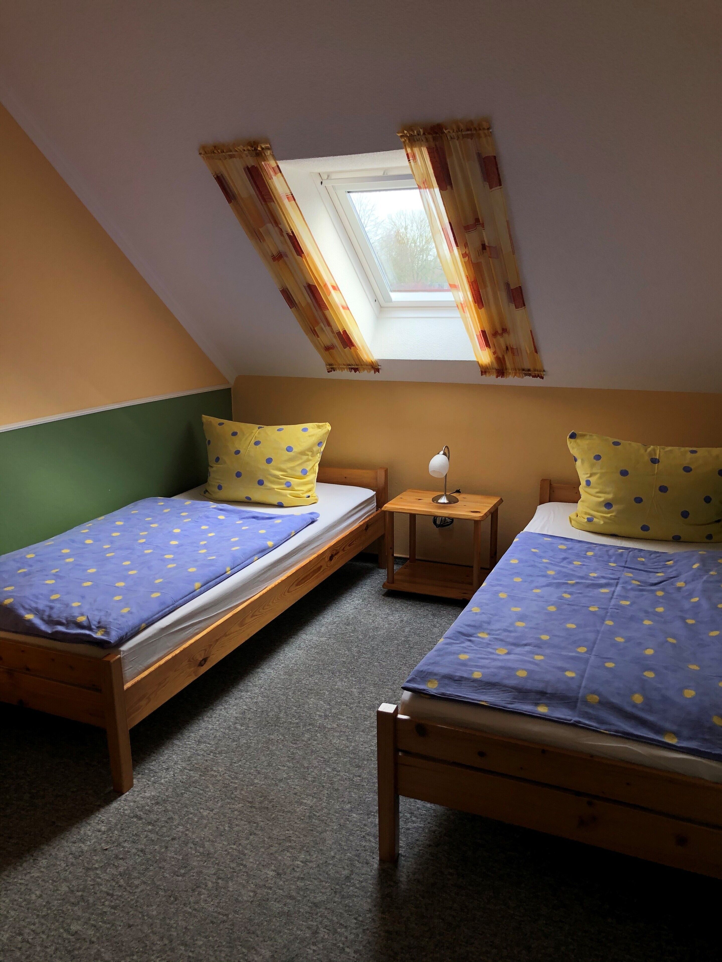 Ein Zimmer mit zwei hölzernen Betten, jeweils mit einer blau-gelben gepunkteten Tagesdecke, einem kleinen Nachttisch mit Lampe und einem Oberlicht mit Vorhängen.
