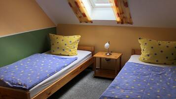 Ein Zimmer mit zwei hölzernen Betten, jeweils mit einer blau-gelben gepunkteten Tagesdecke, einem kleinen Nachttisch mit Lampe und einem Oberlicht mit Vorhängen.