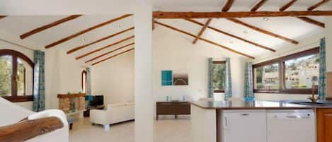Amplia sala de estar con techo alto y vigas de madera a la vista, una cocina con un ventanal grande y vistas del paisaje circundante.
