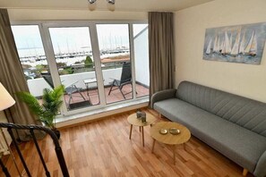 Ein Zimmer mit grauem Sofa, einem kleinen runden Tisch mit zwei Kerzen, einer Zimmerpflanze und Blick auf Segelboote durch große Fenster.