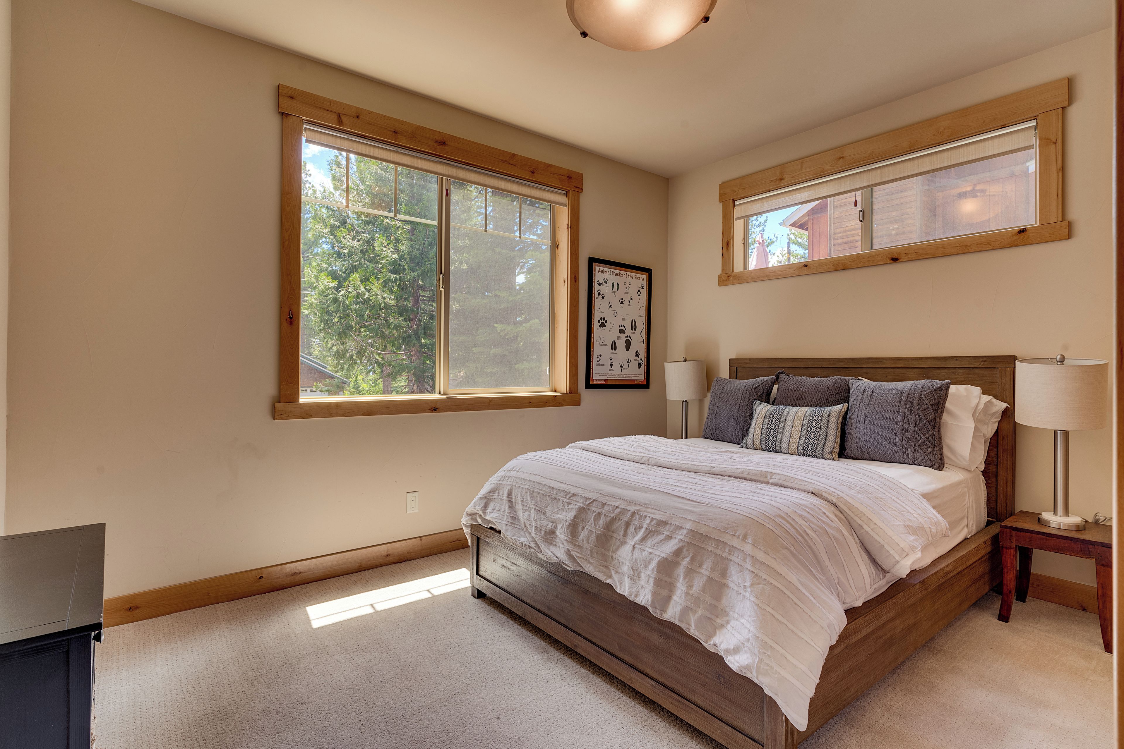 A bedroom with a large bed, two bedside lamps, a framed poster on the wall, and a window with a view of trees.