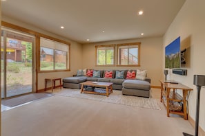 A modern living room with a sectional sofa, a coffee table, and a large window with a view of a wooden deck and trees. - Spacious 5BR Kings Beach Lodge with Mountain Views and Two Living Rooms (Kings Beach)