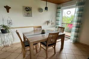 Ein Essbereich mit Holztisch und Stühlen, einer Topfpflanze, einer Uhr und einem Fenster mit Vorhängen.