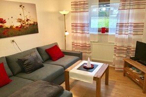 Ein Wohnzimmer mit grauem Sofa, roten Kissen, einem kleinen Tisch mit weißem Tuch, einer Stehlampe und einem Fernseher auf einem Holzständer.