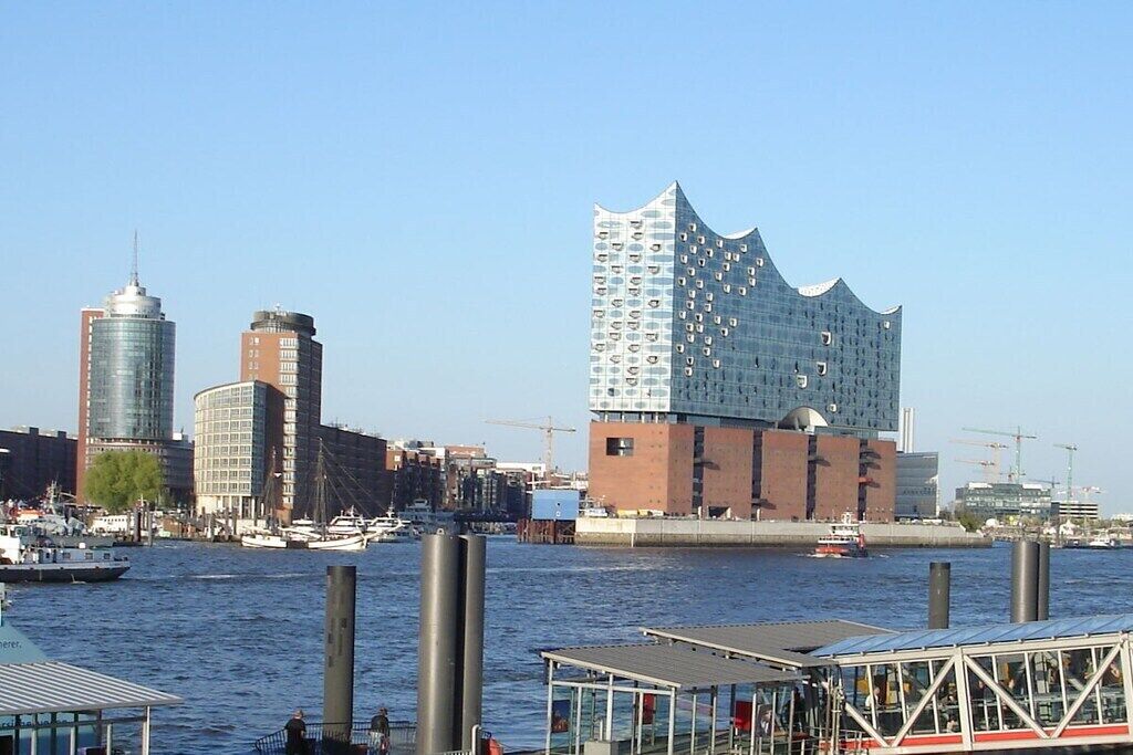 A waterfront cityscape with modern buildings, including a distinctive wavy-roofed structure.