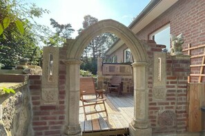 A stone archway leading to a wooden deck with outdoor furniture. - Forest holiday apartment Hamburg 1 (Rosengarten)