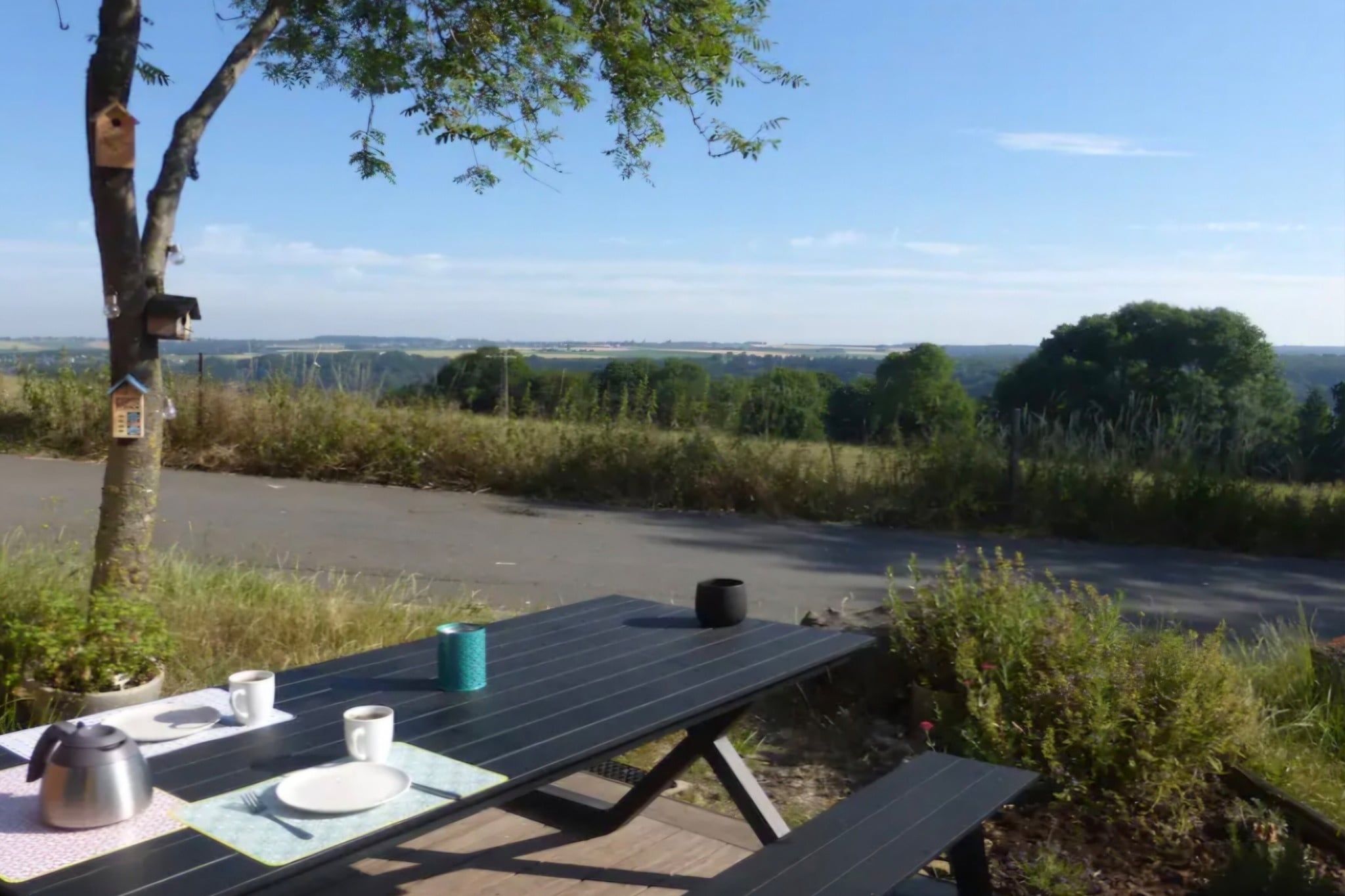 A picnic table with a solar panel, a kettle, and a mug, set against a backdrop of a rural landscape with trees and a clear sky.