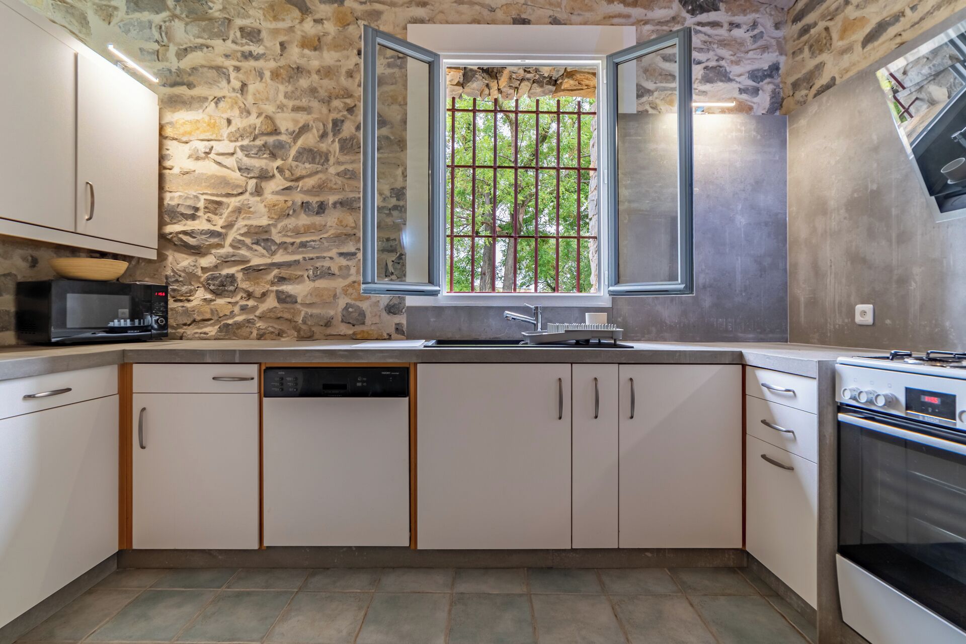 Une cuisine avec des murs en pierre, une fenêtre avec des barreaux, un four à micro-ondes, un lave-vaisselle et une cuisinière.