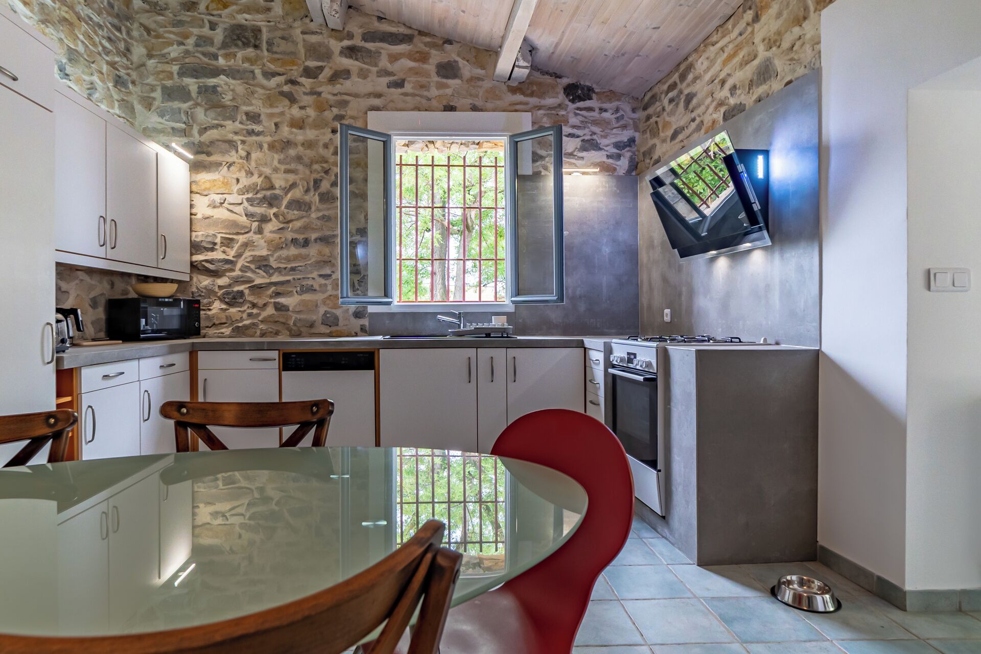A kitchen with stone walls, a dining table with chairs, a TV mounted on the wall, and a window with bars.