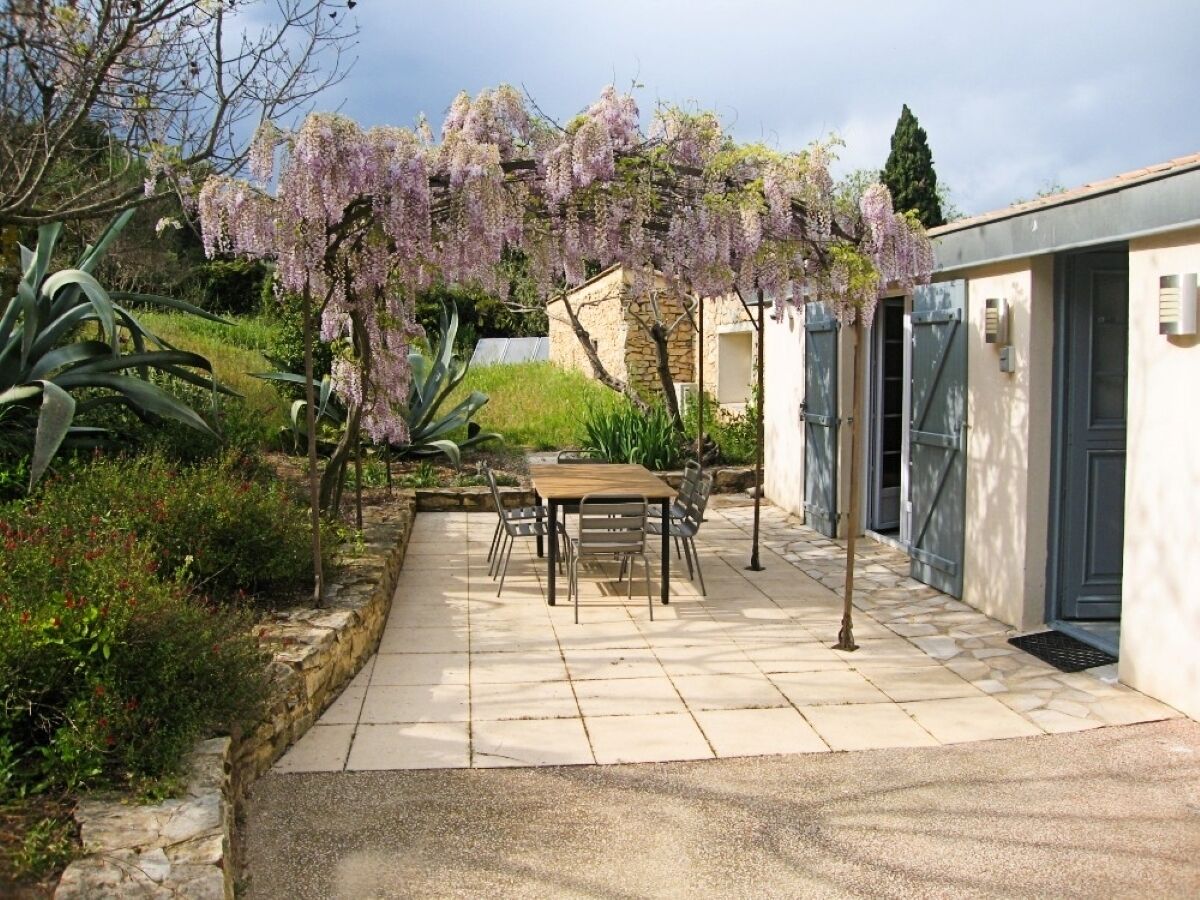 A patio with a table and chairs, surrounded by plants and a building with a door.