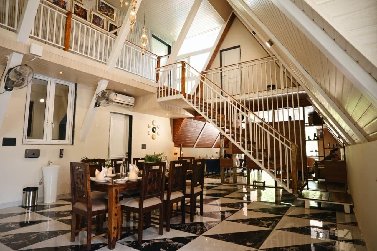 A spacious dining area with a checkered floor, a wooden dining table, and chairs. A staircase with a railing leads to an upper level.