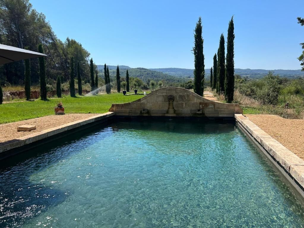 Une piscine aux eaux cristallines, surmontée d’un pont de pierre, le tout entouré de verdure et d’arbres.