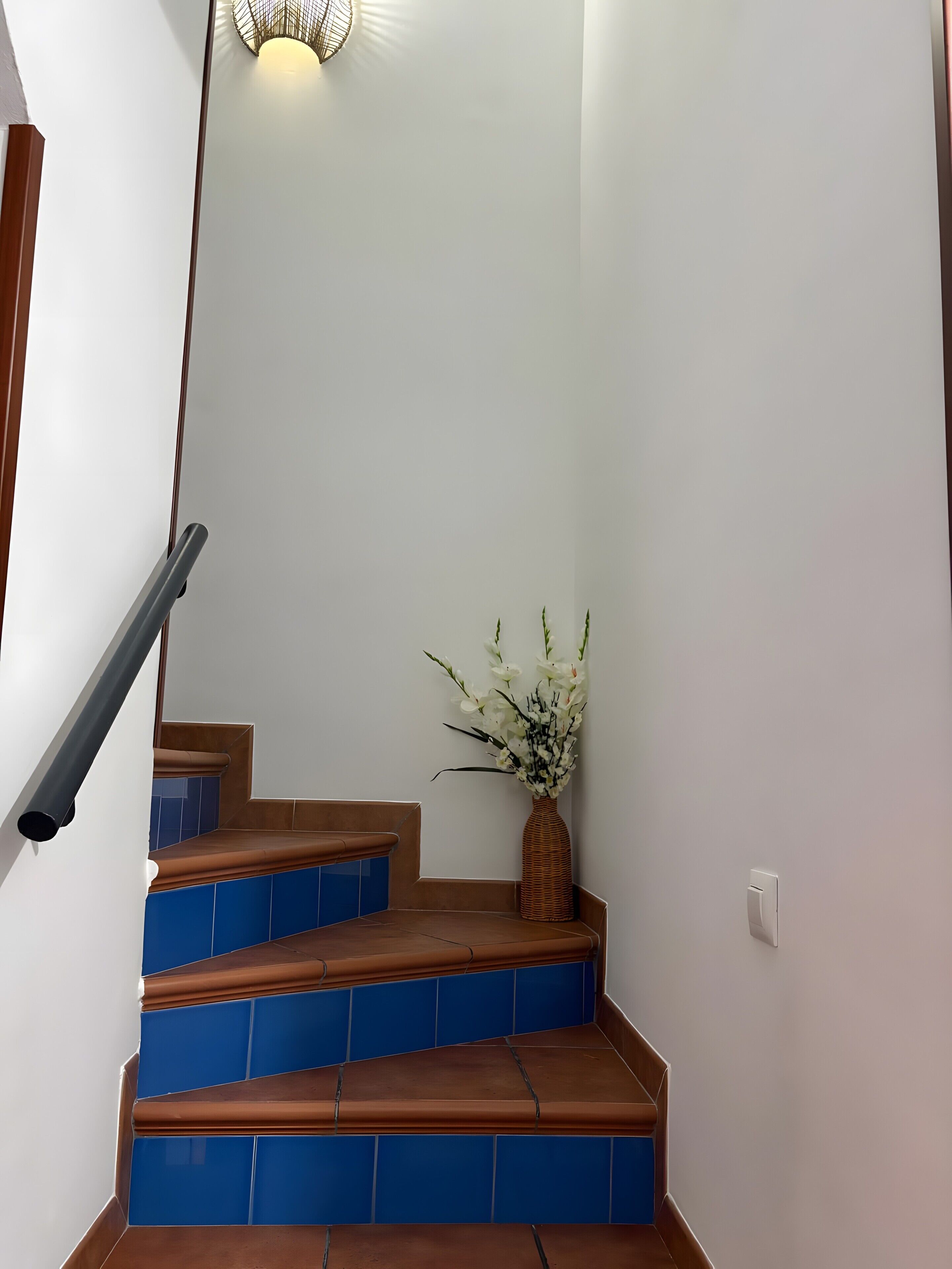 Een trap met blauwe en bruine tegels, een wandlamp en een vaas met witte bloemen.