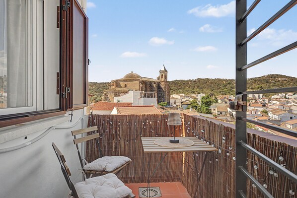 Un balcón con vistas a un edificio histórico con un campanario, una mesa con una lámpara y una silla.