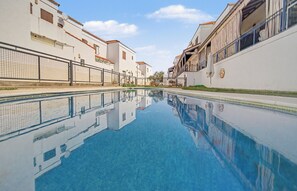 Una piscina con superficie de agua azul cristalina, rodeada de edificios blancos con balcones y una cerca.