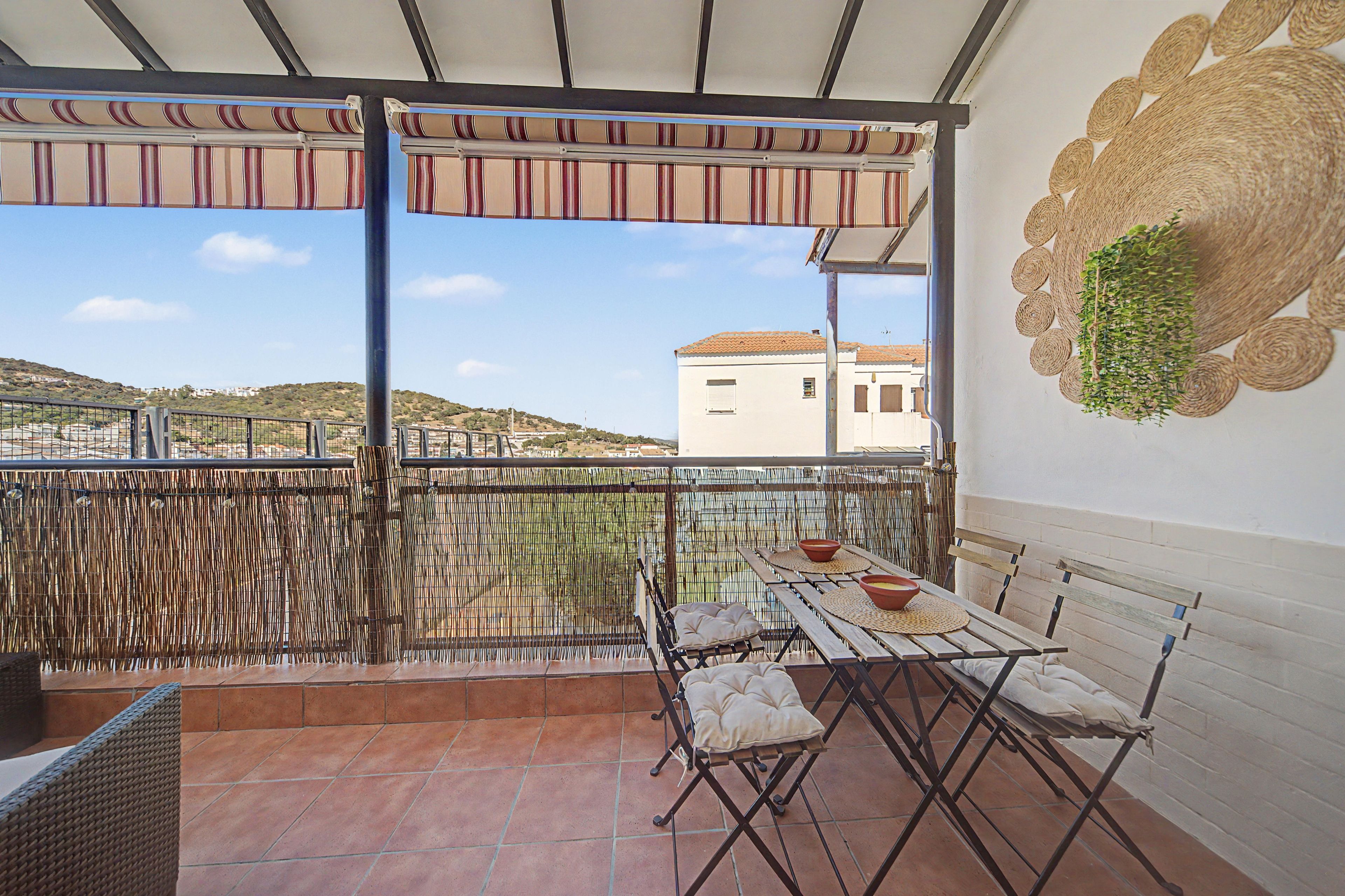 Un balcón con mesa y sillas, un cerramiento tejido y vistas a una colina y edificios.