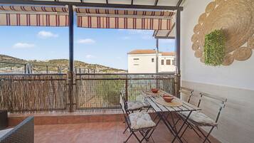 Un balcón con mesa y sillas, un cerramiento tejido y vistas a una colina y edificios.