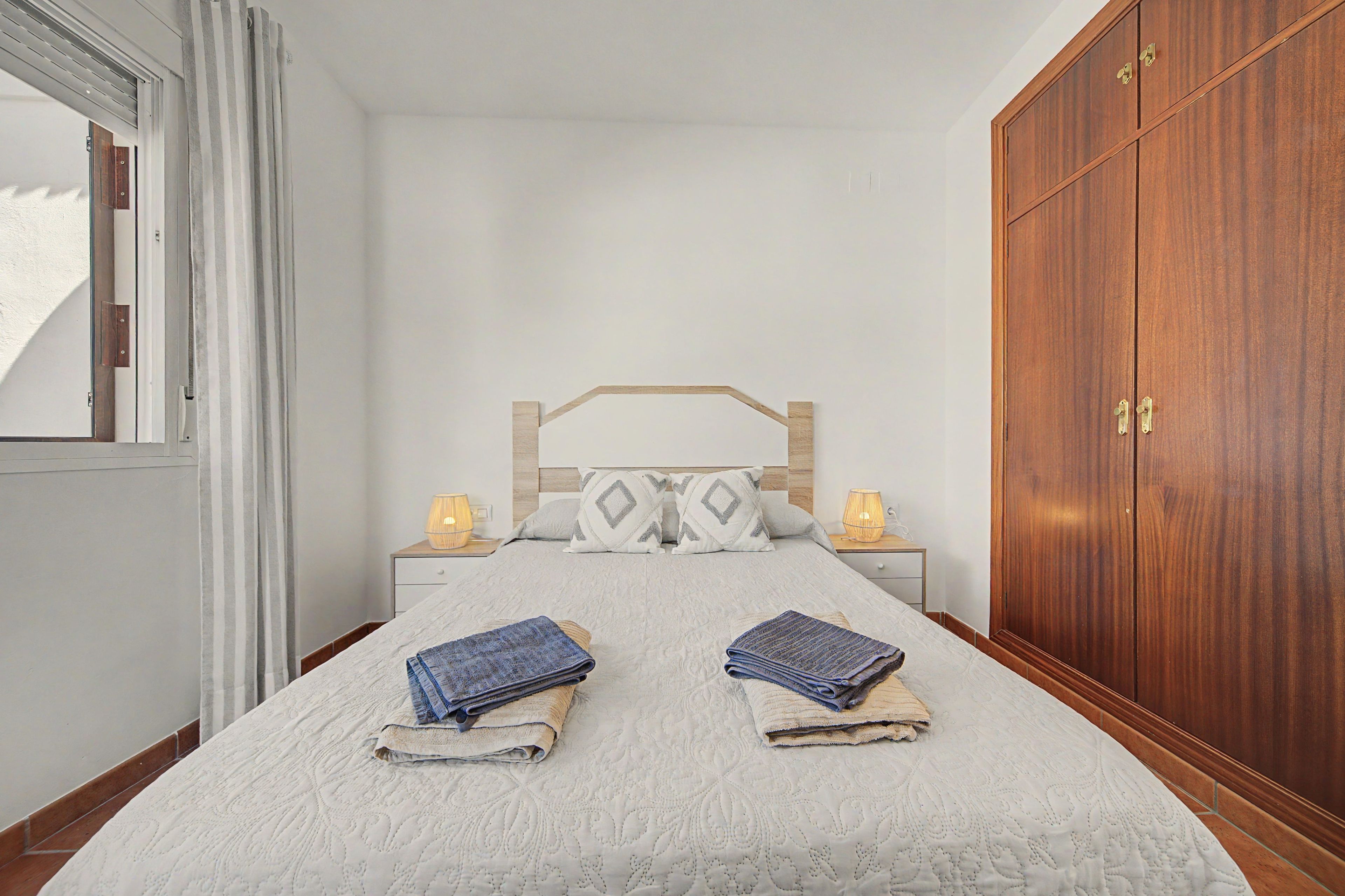 Un dormitorio con cama, lámparas de noche, armario y ventana con cortinas.