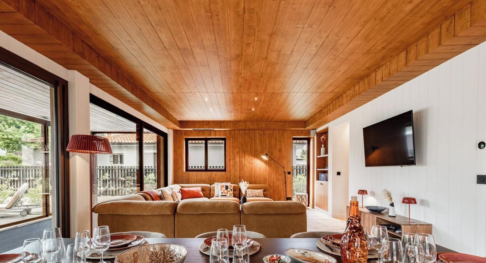 Et moderne interiør med træloft, en sofa, et spisebord med glas og tallerkener, og et fjernsyn ophængt på væggen.