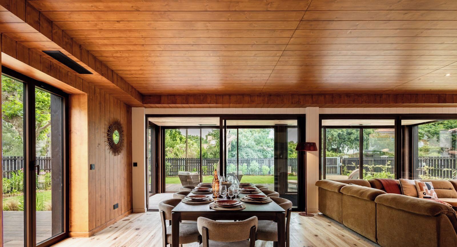 A dining area with a wooden ceiling, large windows, and a sofa.